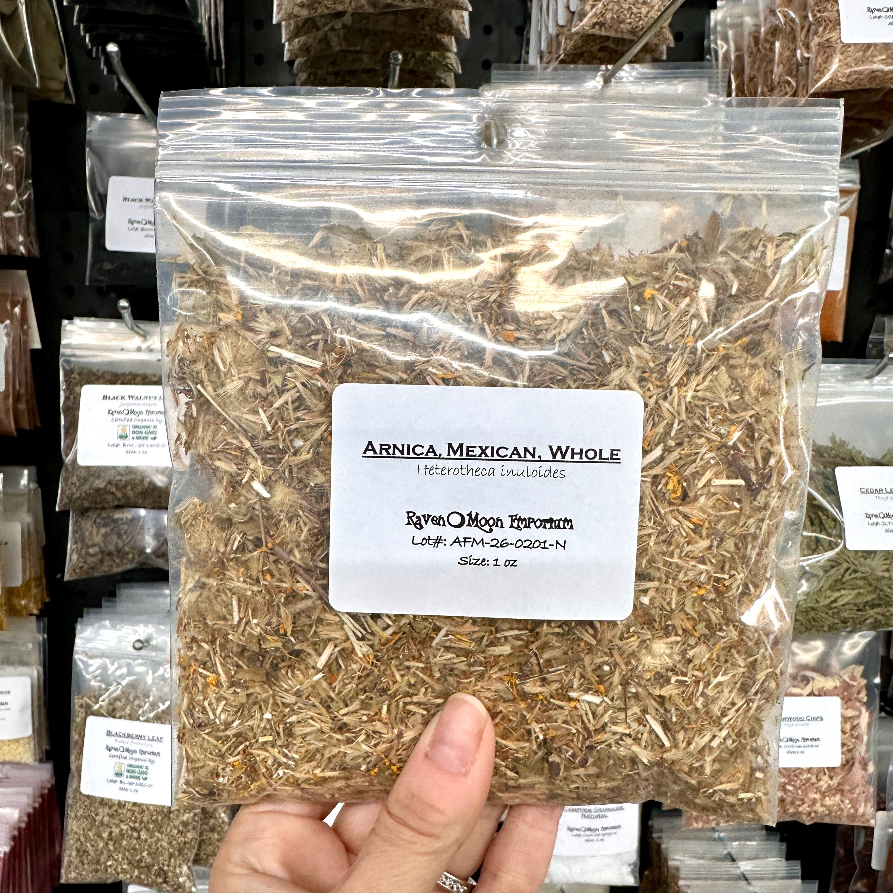 A hand holds a clear plastic zip bag filled with dried Mexican Arnica Whole Flowers. The bag is labeled with the Raven Moon Emporium logo.