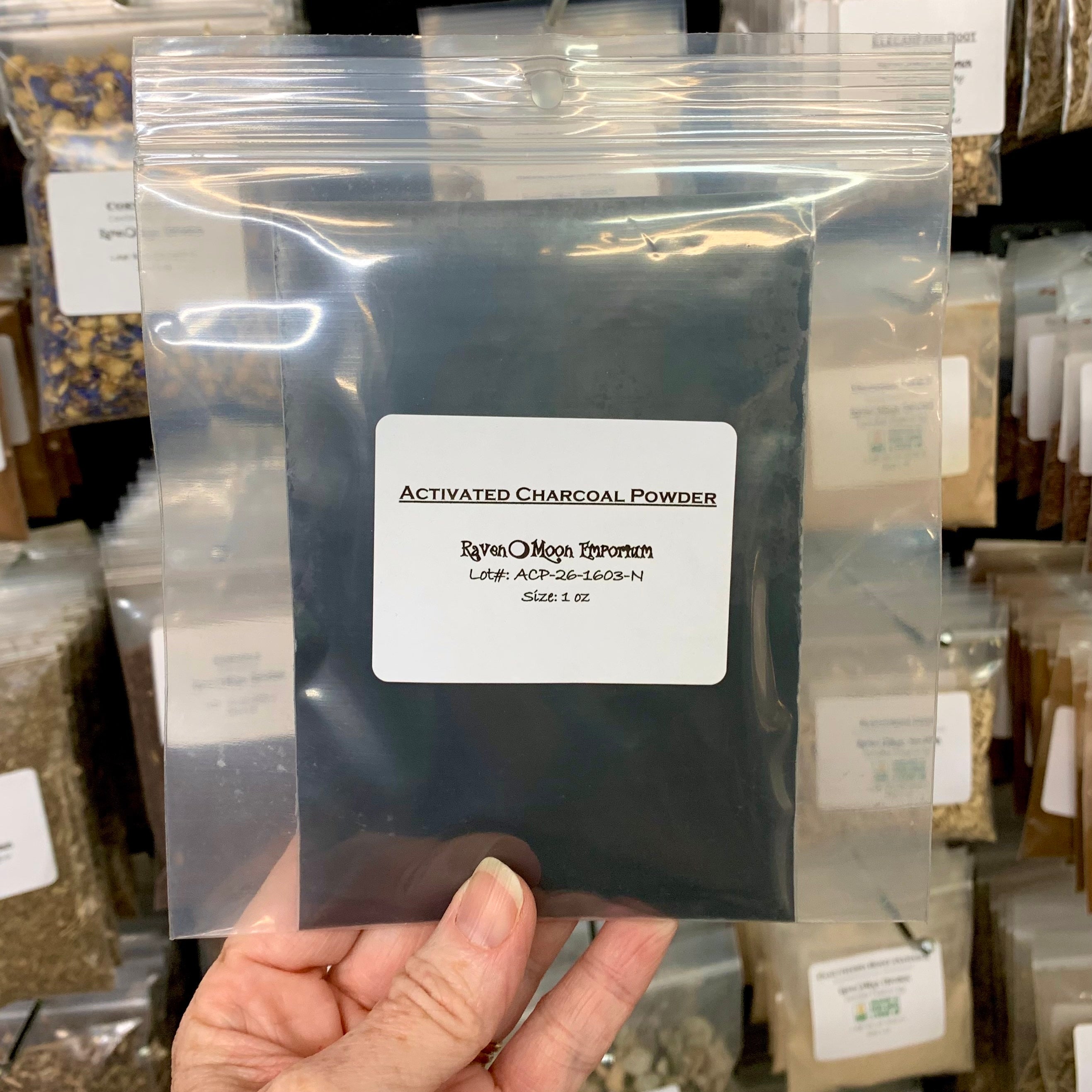 Hand holding a double-bagged, clear zip bag of activated charcoal powder. The bag is labeled with The Raven Moon Emporium logo.