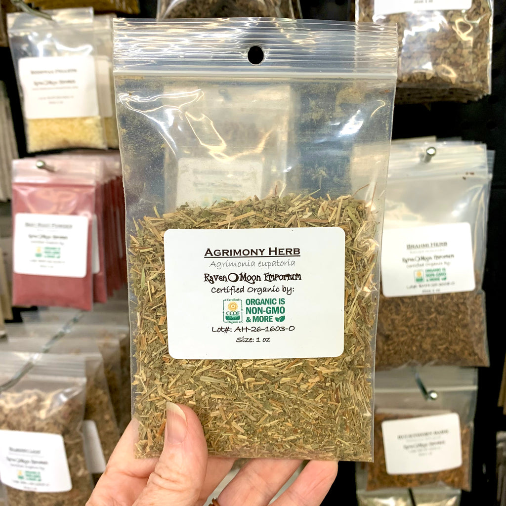 Person's hand holding  a one-ounce clear plastic zip bag of packaged agrimony herb labelled with the Raven Moon Emporium logo. In the background are other similarly packaged herbs.