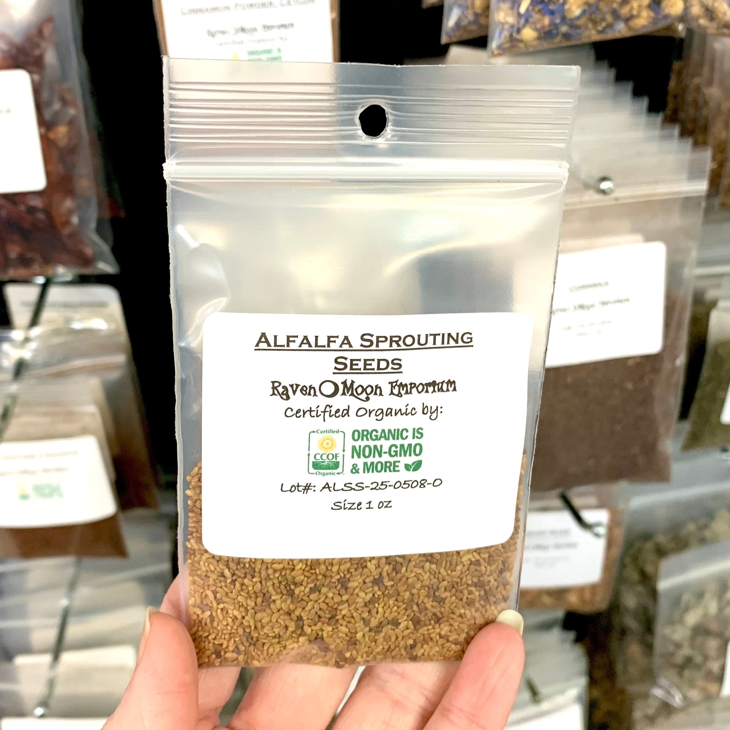 Hand holding a clear zip bag of alfalfa sprouting seeds with a background of similar packages. The bag is labeled with the Raven Moon Emporium logo.