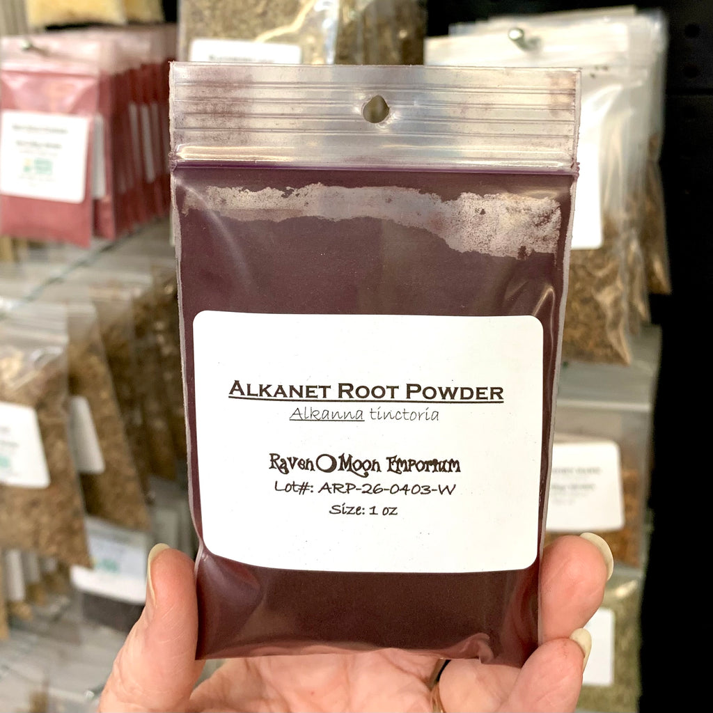Hand holding a clear zip bag of Alkanet Root Powder in front of several similar herb packages. The bag is labeled with the Raven Moon Emporium logo.