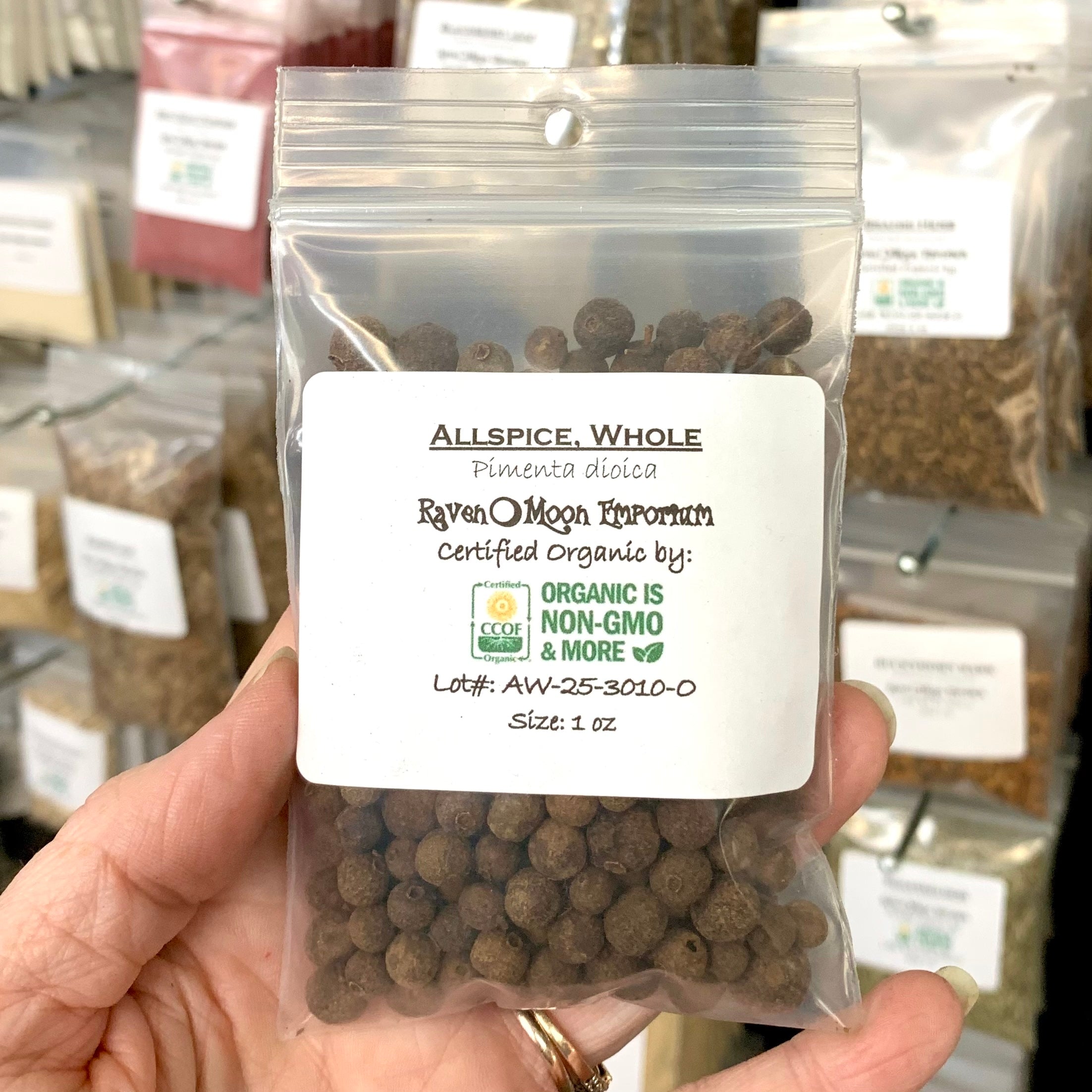 Hand holding a clear zip bag of organic whole allspice with other similar bags of herbs in the background. The bag is labeled with the Raven Moon Emporium logo.