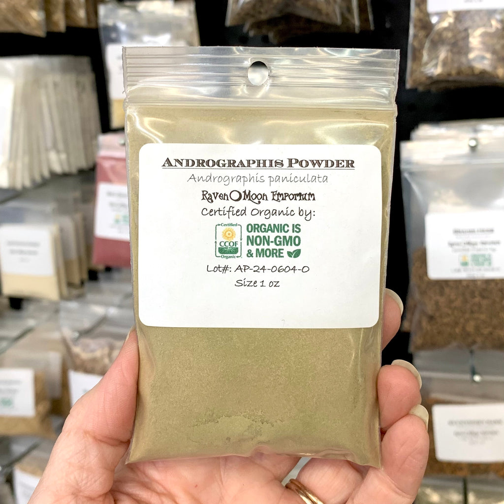 Hand holding a clear zip bag package of Andrographis Powder. The bag is labeled with the Raven Moon Emporium logo.