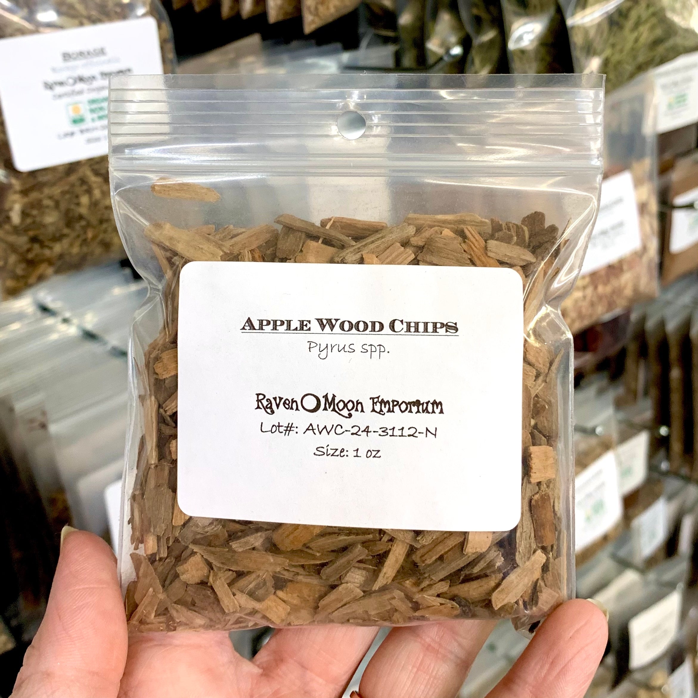 Hand holding a clear zip bag of apple wood chips. The bag is labeled with the Raven Moon Emporium logo.