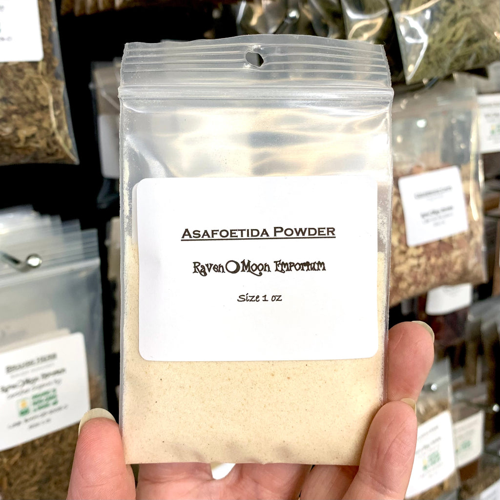 Hand holding asafoetida powder in a clear zip plastic bag. The bag is labeled with the Raven Moon Emporium logo.