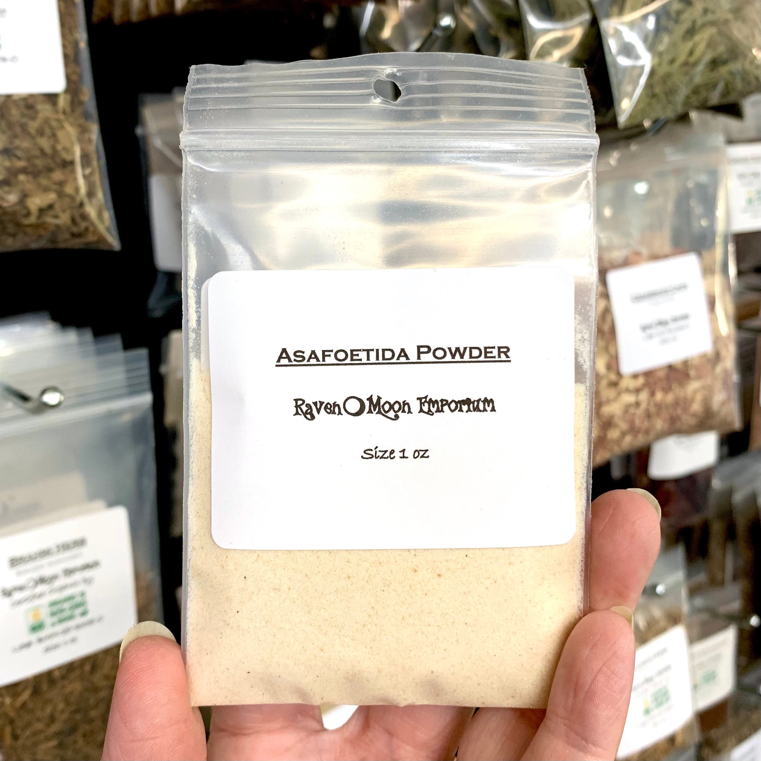 Hand holding asafoetida powder in a clear zip plastic bag. The bag is labeled with the Raven Moon Emporium logo.