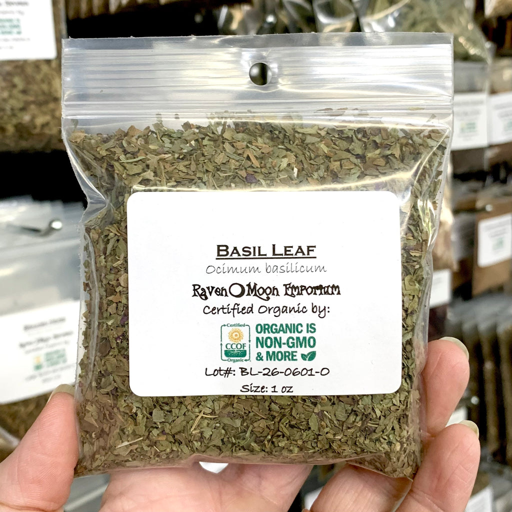 A hand holds a clear plastic zip bag of dried basil leaf pieces. The bag is labeled with the Raven Moon Emporium logo.