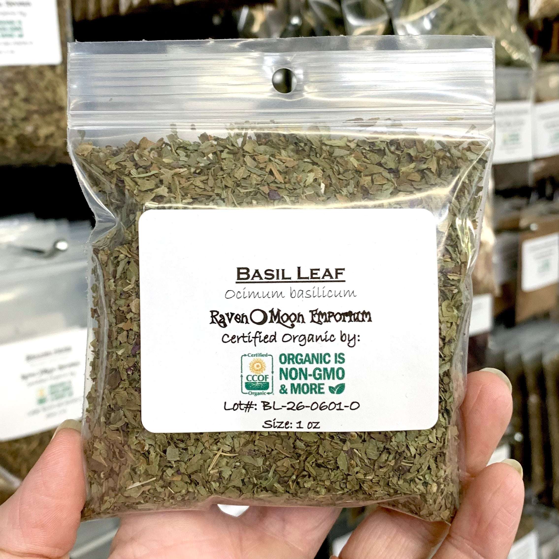 A hand holds a clear plastic zip bag of dried basil leaf pieces. The bag is labeled with the Raven Moon Emporium logo.
