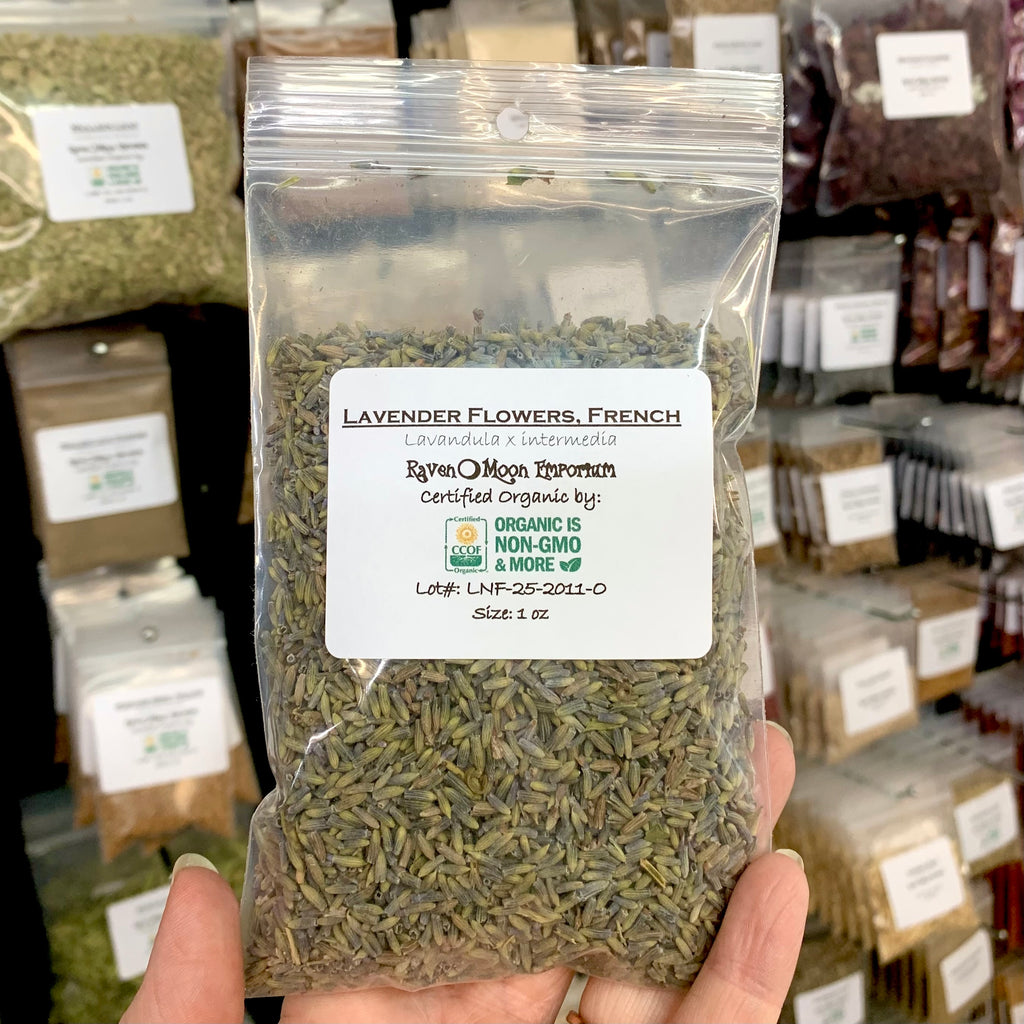 Hand holding a clear plastic zip bag of dried French lavender flower buds with a blurred background of similar packages. The bag is labeled with the Raven Moon Emporium logo.