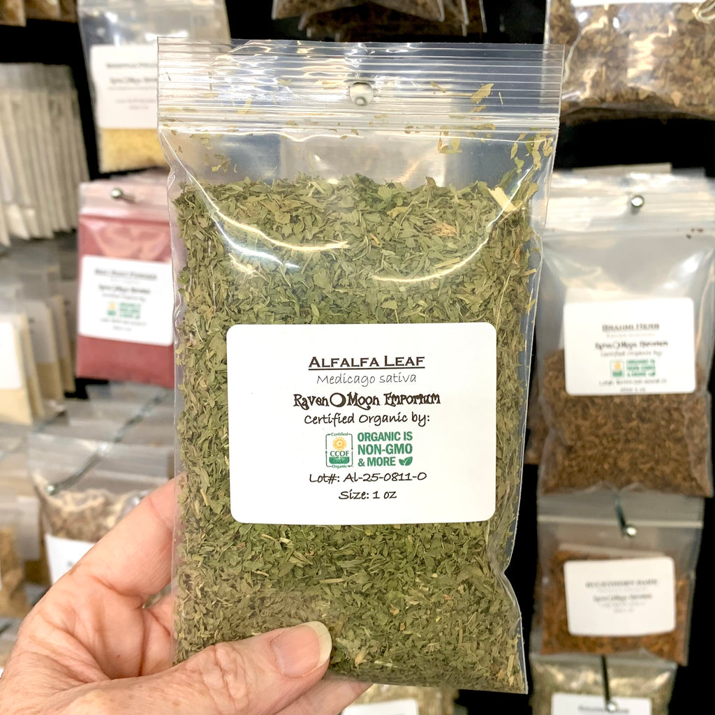 Hand holding a clear zip bag of dried, cut alfalfa leaf. The bag is labeled with the Raven Moon Emporium logo.