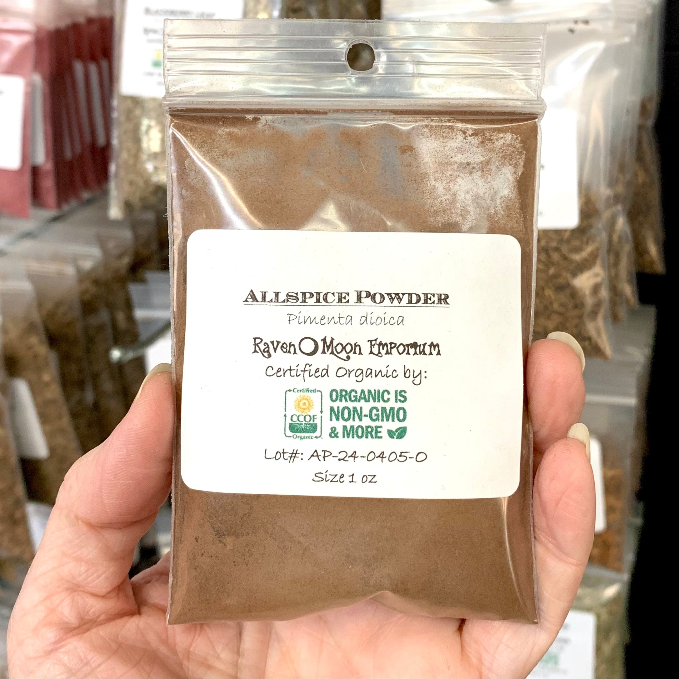 Hand holding a clear plastic zip bag of allspice powder with a label in front of a background of similar bags of herbs. The bag is labeled with the Raven Moon Emporium logo.