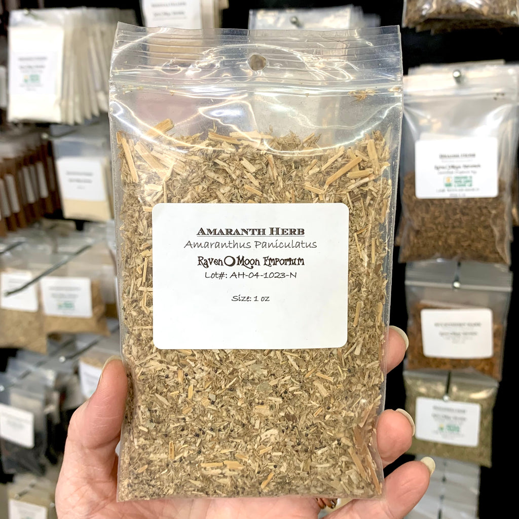 Hand holding a clear zip bag of amaranth herb with other similar bags of herbs in the background. The bag is labeled with the Raven Moon Emporium logo.